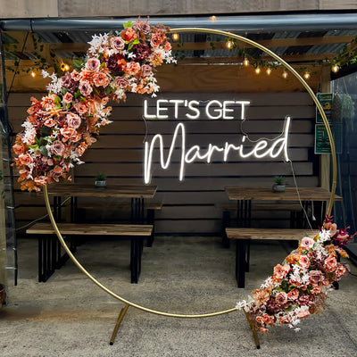 white lets get married neon sign hanging in gold hoop backdrop with flowers