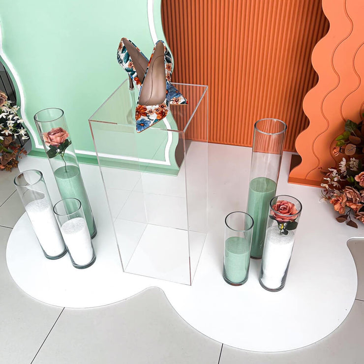clear plinth standing on white mat with heels on top and green and white sand vases on side