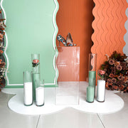 clear plinth standing on white floor mat in front of green and orange backdrop boards and coloured sand candles