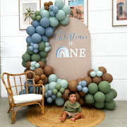 one year old boy sitting on round floor rug in front of arch backdrop with balloons and arm chair