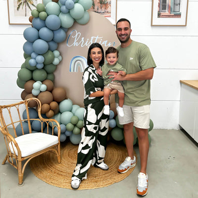 family with baby standing in front of brown arch backdrop with balloons, arm chair and rug