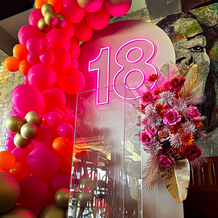 hot pink 18 neon sign hanging on arch backdrop with balloon garland, flower arrangement and clear plinth