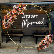 white lets get married neon sign hanging on gold hoop backdrop with flower arrangement
