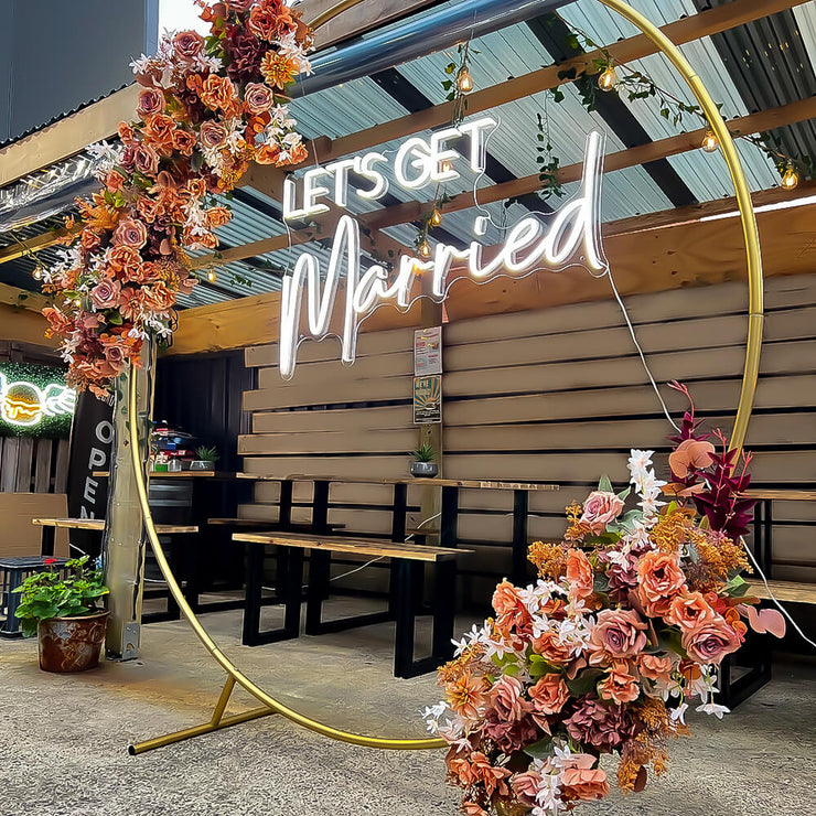 white lets get married neon sign hanging on gold hoop backdrop with artificial flower garland