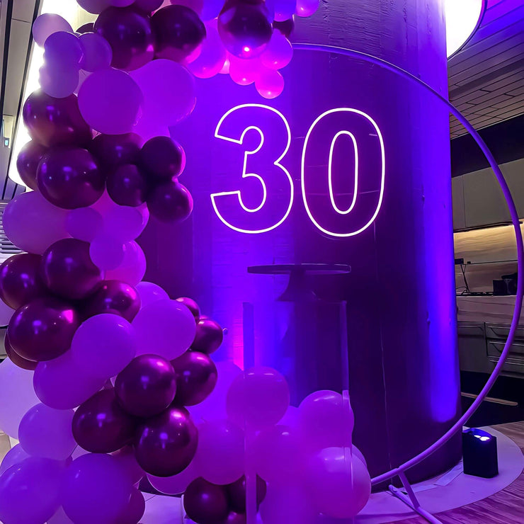 purple 30 neon sign hanging on white hoop backdrop with balloons and clear plinth