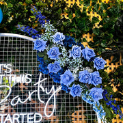blue rose and baby's breath flower arrangement hanging on white arch backdrop frame against green garden wall
