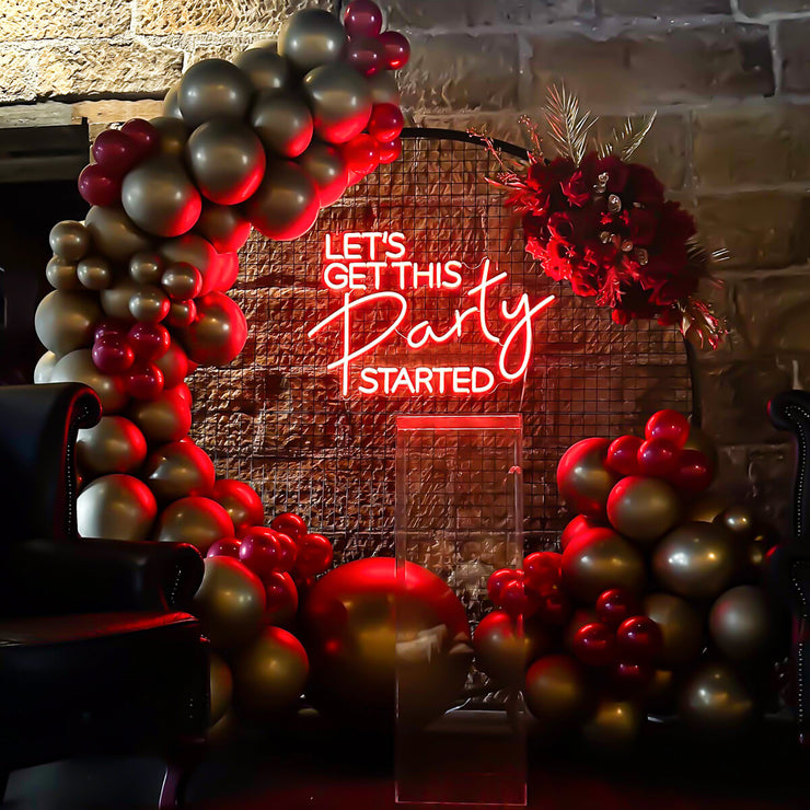 red lets get this party started neon sign hanging on black circle backdrop with gold and red balloons and flowers