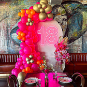pink 18 neon sign hanging on beige arch backdrop with pink and gold balloons and flowers