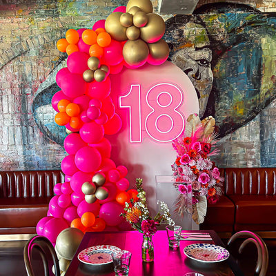 pink 18 neon sign hanging on beige arch backdrop with pink and gold balloons and flowers