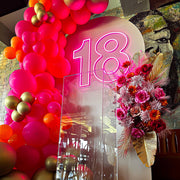 pink 18 neon sign hanging on beige arch backdrop with flowers, balloons and clear plinth