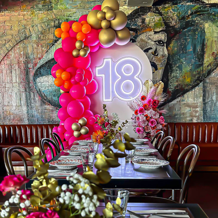 white 18 neon sign hanging on beige arch backdrop with pink balloons and flowers at restaurant