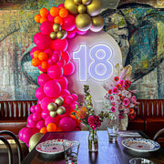white 18 neon sign attached to beige arch backdrop with pink and gold balloon arch and flower arrangement