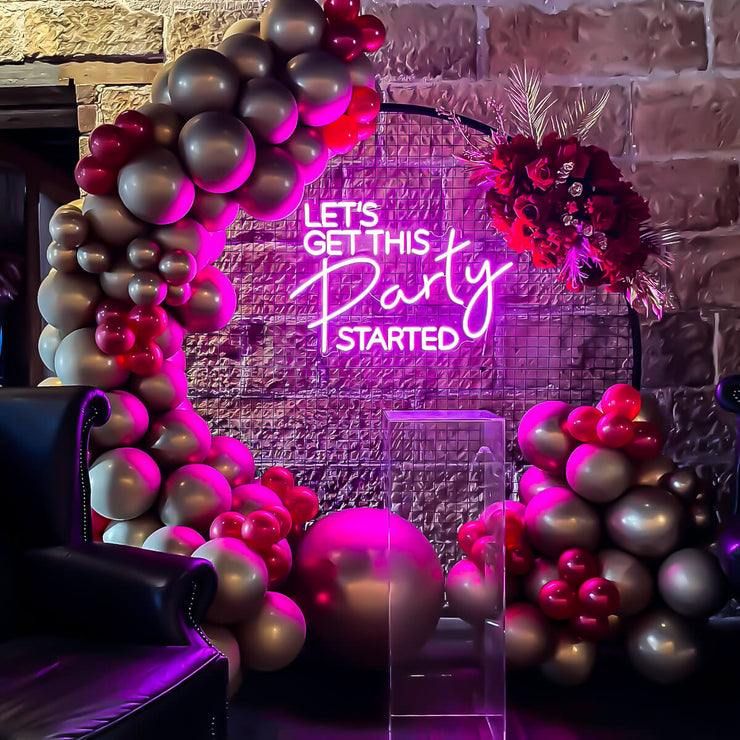 pink lets get this party started neon sign hanging on circle mesh backdrop with balloons, plinth and flower arrangement