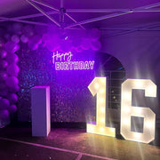 purple happy birthday neon sign hanging on wall in front of big stand up numbers and balloons