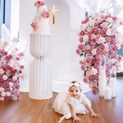 baby sitting on floor in front of white slatted plinth and pink and lilac rose flower arrangements