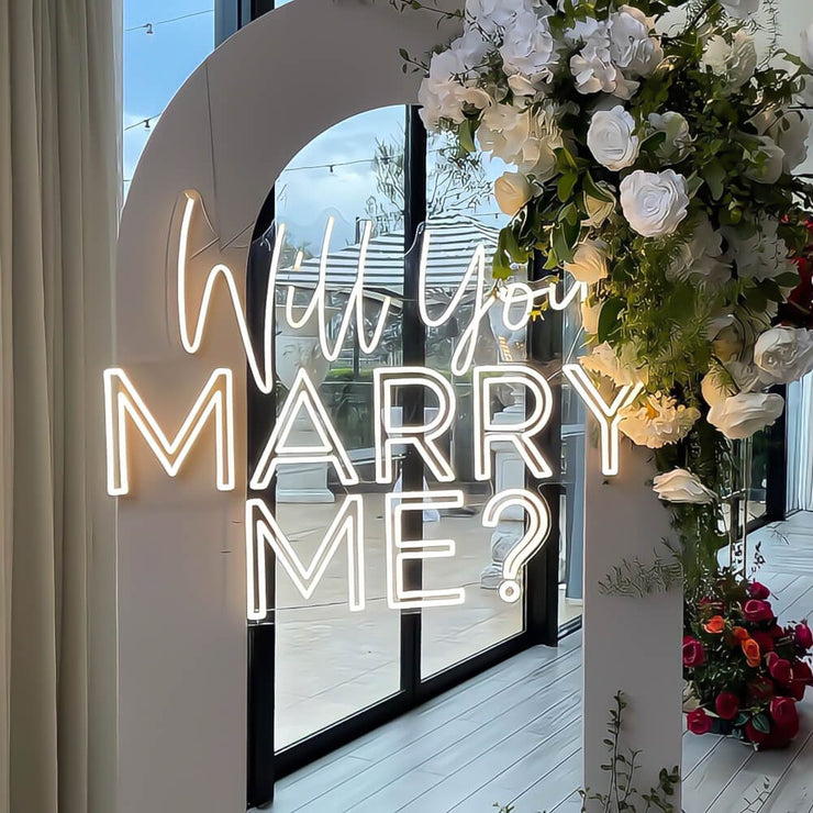 warm white will you marry me neon sign hanging on arch backdrop with flowers at wedding